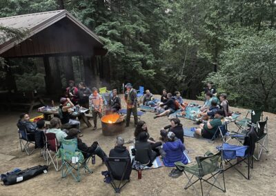 25 Trans Campers sitting on blankets or camp chairs around the campfire in the daytime in a forest