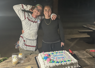 Two people with arms around each other facing the camera behind a cake with words "Welcome to Camp"